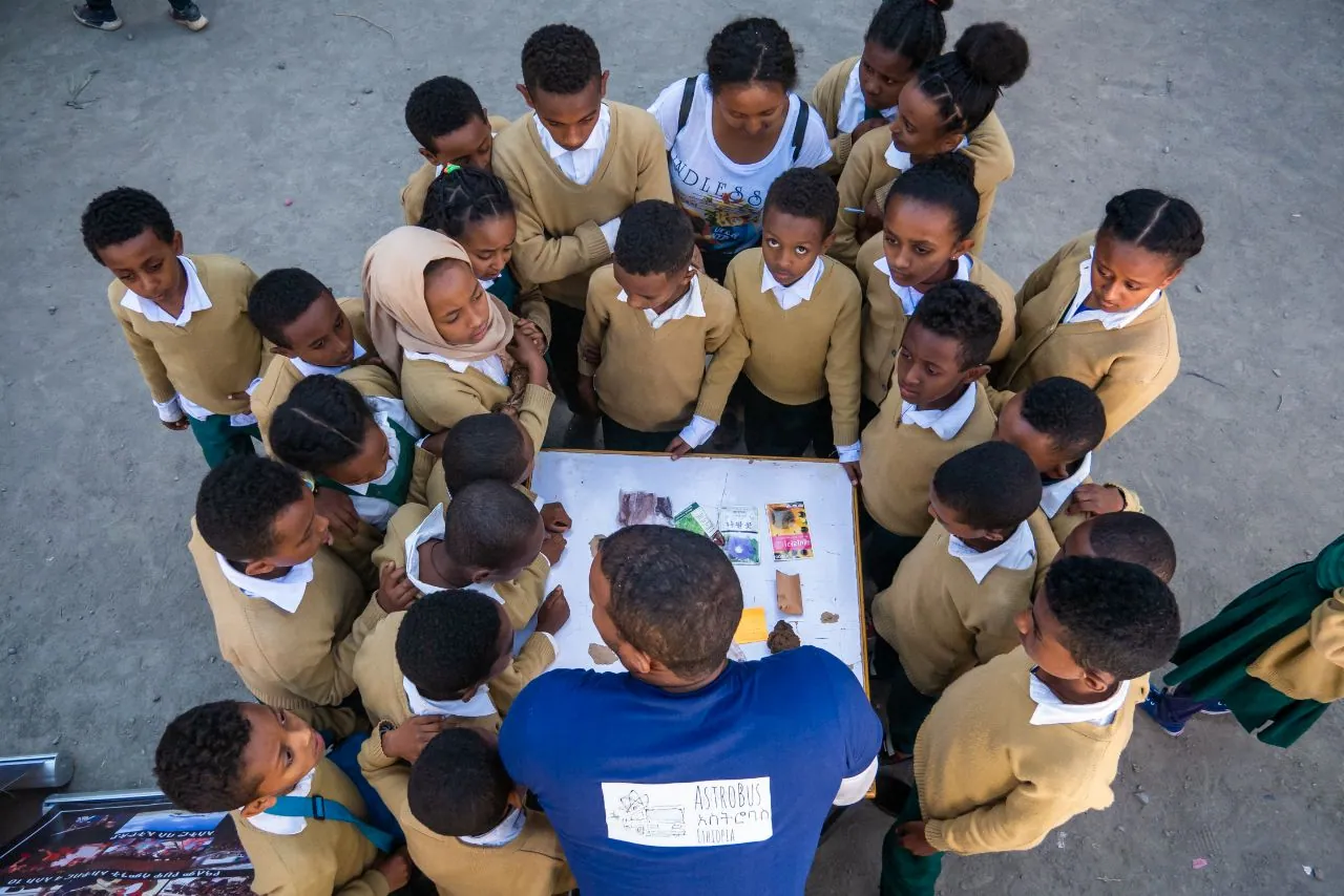 Seen from above, students crowd round a team member and a table of pictures