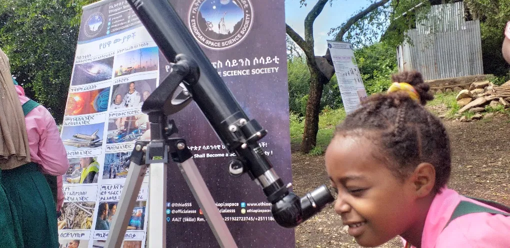 A girl looks through a telescope beside the Society's banner