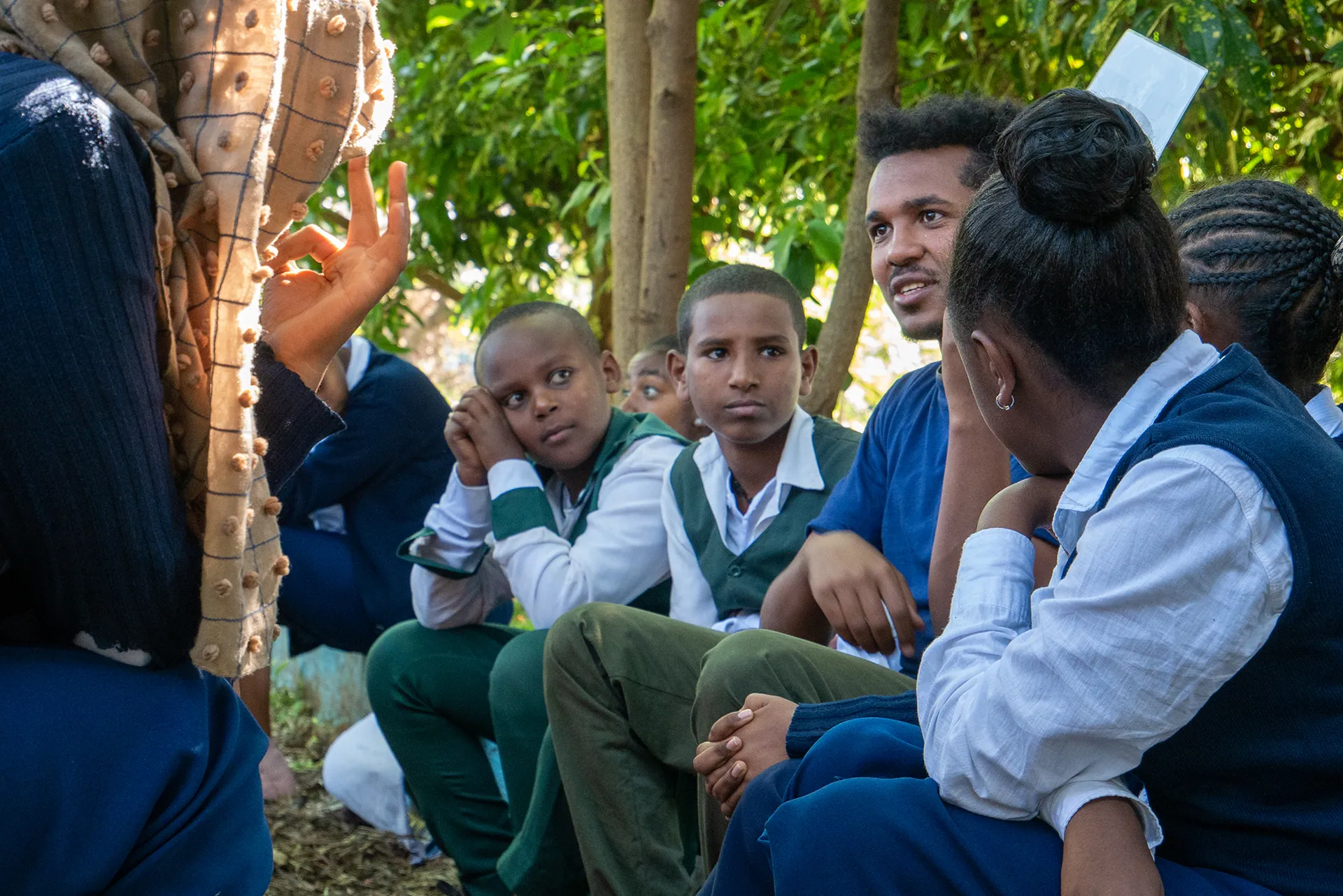 A team member sits with students under the trees, listening